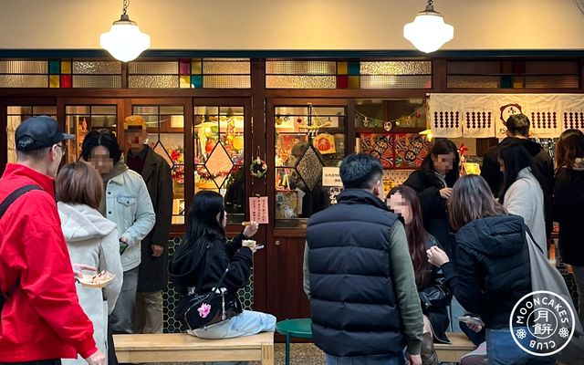 A crowd eats and waits in front of many sets of sliding dark wooden doors decorated with stained and patterned glass.