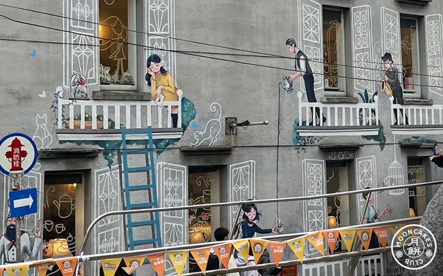 A cement wall with windows, each of which have painted on them a balcony with fancy window shades and people doing various things like playing saxaphone and pouring coffee.